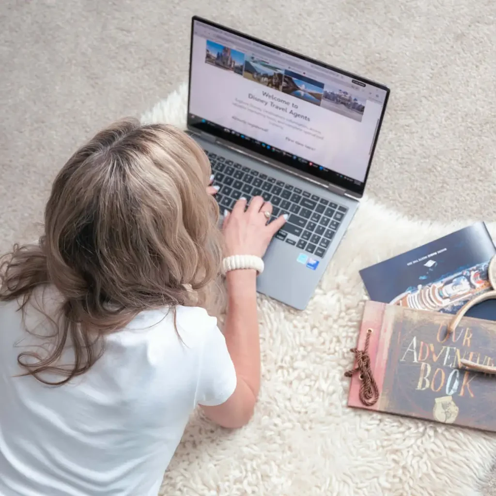 A Travel Agent with wavy blonde hair types on a laptop while sitting on a fluffy rug in her family room. Nearby are a blue passport, a pen, and a book titled Our Adventure Book. - Favorite Grampy Travels