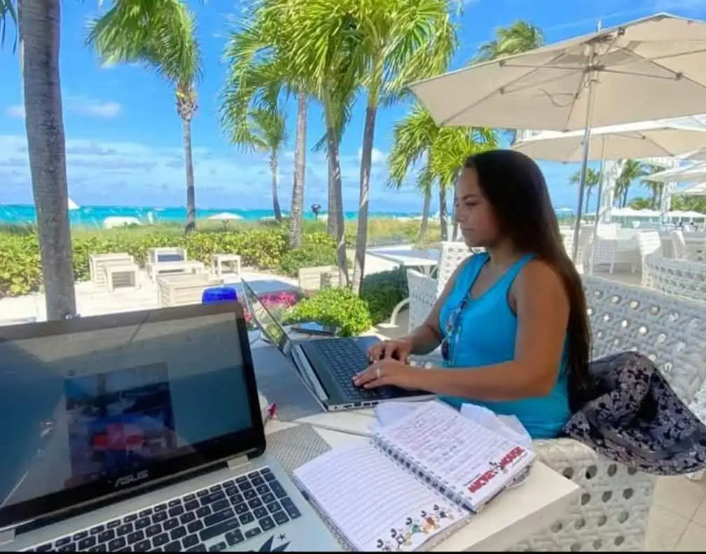 Travel Agent working on a laptop at an outdoor beachside café with palm trees, a notebook, and another open laptop nearby; the ocean and blue sky are in the background. - Favorite Grampy Travels