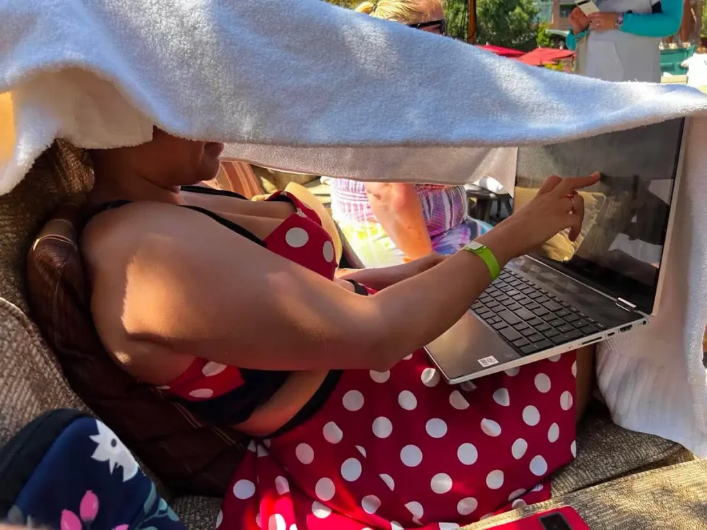 A Travel Agent in a red polka dot swimsuit uses a laptop outdoors, shielding the screen with a towel draped overhead to block sunlight. - Favorite Grampy Travels