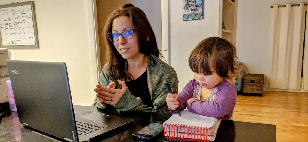 A woman wearing glasses works on a laptop while a toddler sits beside her, drawing in a notebook with a pen. A phone and notebook are on the table. They appear to be in a home kitchen or dining area. - Favorite Grampy Travels