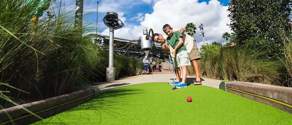 Two people play mini golf outside on a bright, sunny day at Universal CityWalk Orlando, surrounded by tall grass and greenery. One person is putting a red golf ball on a green artificial turf course, making it a perfect outing for anyone looking for inspiration in their Family Guide. - Favorite Grampy Travels