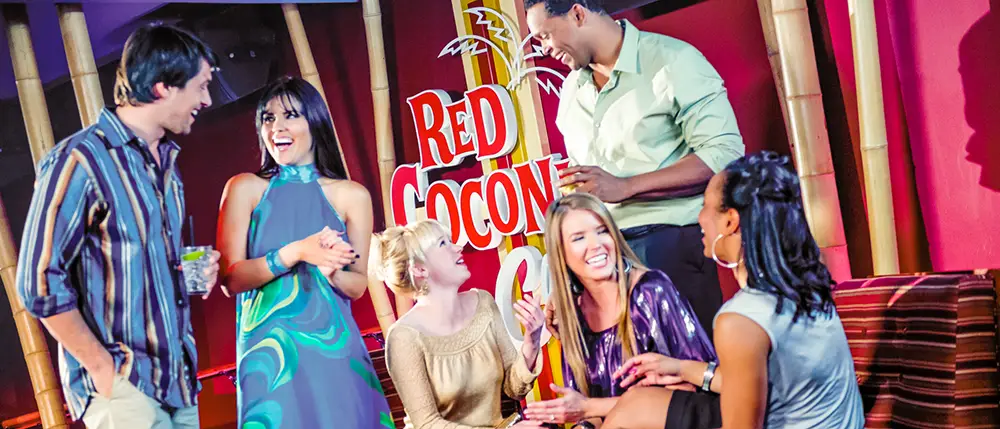 Six people socializing and laughing at a lounge with a colorful Red Coconut sign in the background at Universal CityWalk Orlando. Some are standing, some are sitting, and drinks are in hand, capturing a vibrant moment perfect for any family guide to fun nightlife spots. - Favorite Grampy Travels