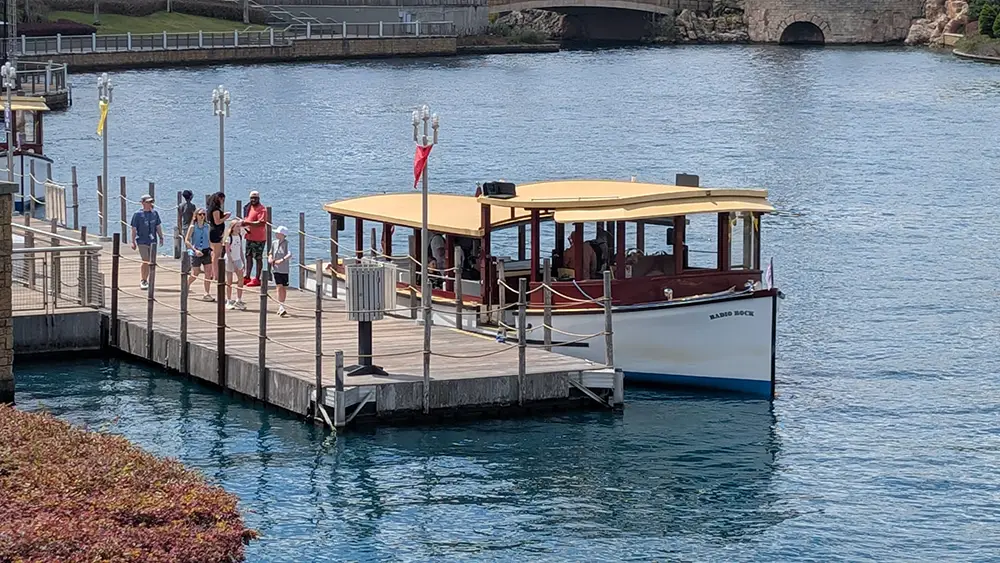 A small boat named Island Rock is docked at a wooden pier as several people stand nearby, capturing a scene reminiscent of the tranquil moments you might find in a Family Guide to Universal CityWalk Orlando. There is a bridge and greenery in the background, and the water is calm and blue. - Favorite Grampy Travels