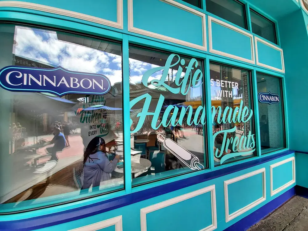 Exterior of a Cinnabon storefront at Universal CityWalk with teal trim and large window decals that read, Life is better with handmade treats. - Favorite Grampy Travels