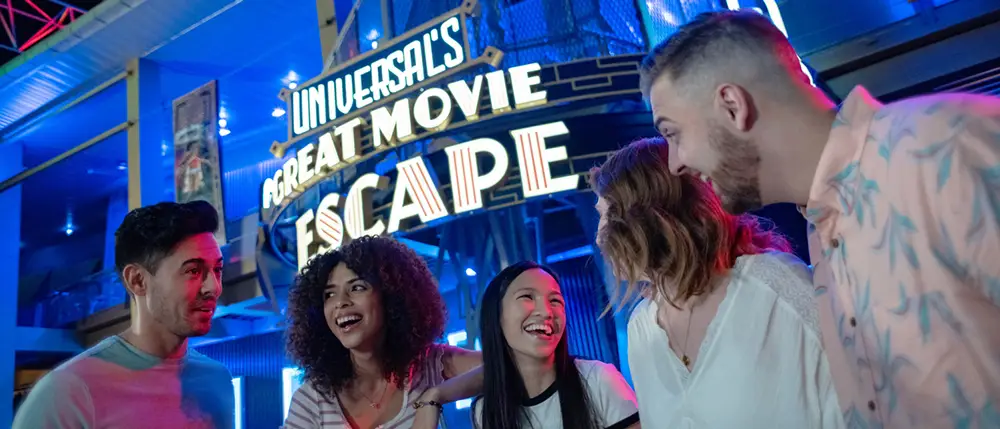 Four people smile and laugh together at night in front of a brightly lit Universals Great Movie Escape sign at Universal CityWalk Orlando, suggesting an energetic and fun atmosphere perfect for any Family Guide. - Favorite Grampy Travels