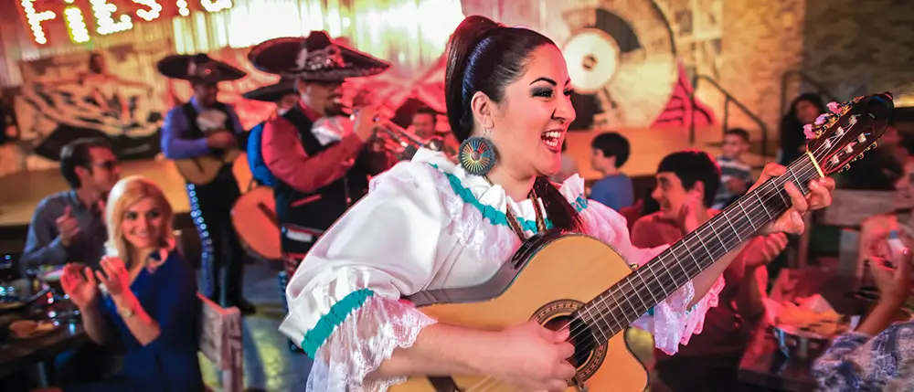A smiling woman in traditional dress plays guitar at a lively indoor celebration at Universal CityWalk Orlando, with musicians in sombreros and people clapping and enjoying the festive atmosphere in the background—a perfect scene for any family guide exploring vibrant cultural experiences. - Favorite Grampy Travels