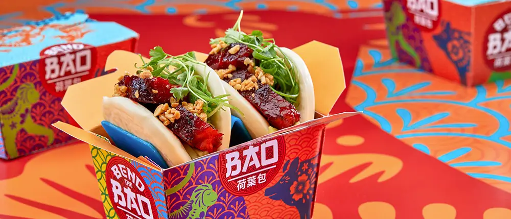 Two bao buns filled with crispy pork, greens, and crushed peanuts are served in a colorful, patterned takeout box at Universal CityWalk Orlando. Similar boxes appear in the background on a vibrant, floral-patterned table—a scene perfect for any Family Guide exploring delicious bites together. - Favorite Grampy Travels