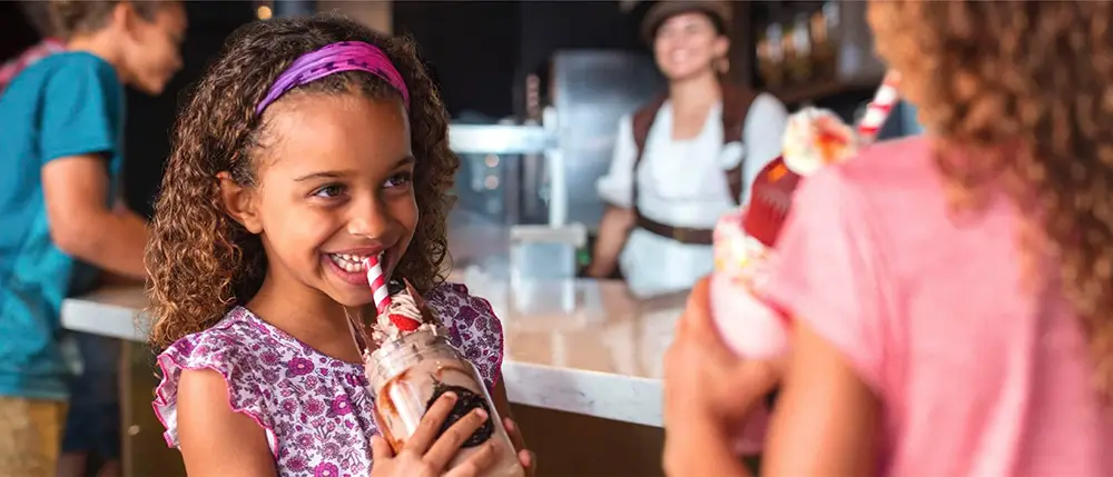 Smiling young girl with curly hair drinks a chocolate milkshake with whipped cream and a striped straw in a restaurant at Universal CityWalk Orlando, while another child holds a similar drink nearby. A server is blurred in the background, capturing a joyful moment perfect for any Family Guide. - Favorite Grampy Travels