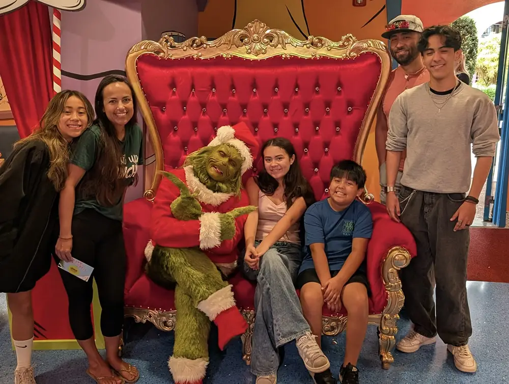 A group of six people poses for a photo with The Grinch, who sits on a large, ornate red velvet throne during Grinchmas at Universal Orlando
