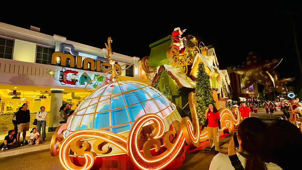 A Santa Claus float with a lit globe and golden scrollwork rolls past the Minion Cafe at night during the holiday parade at Universal Studios Florida