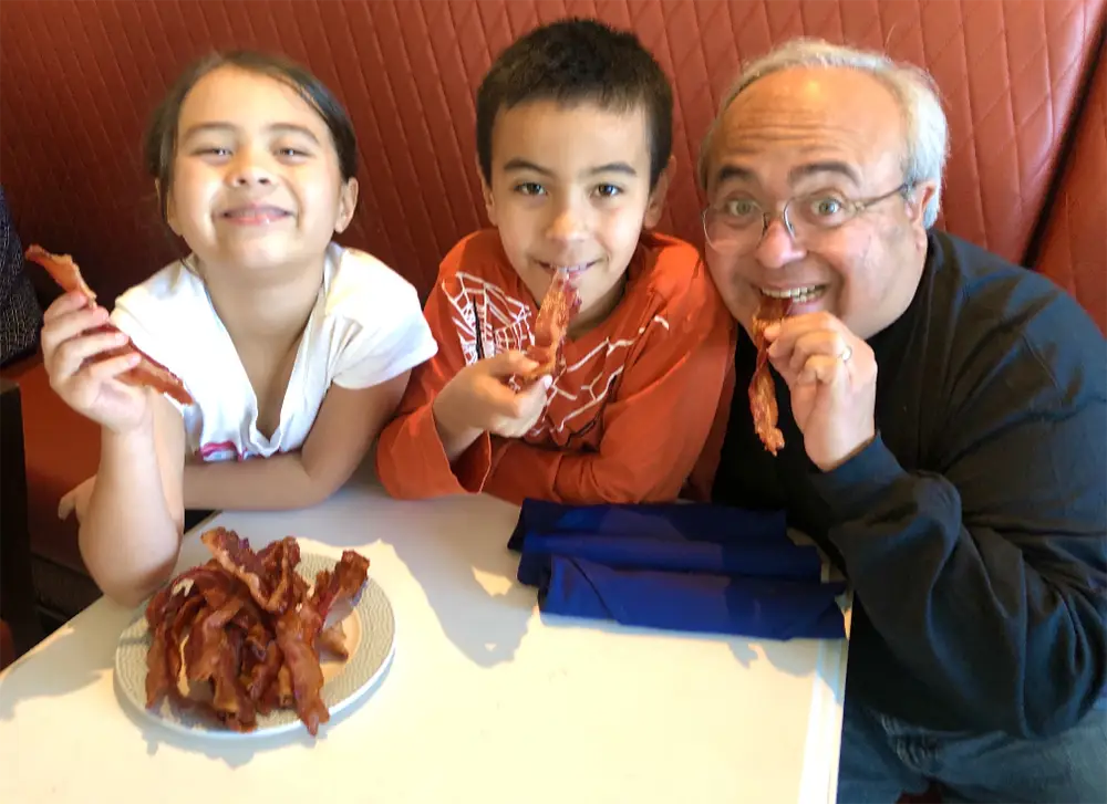 A girl, a boy, and an older man eating strips of bacon together at a restaurant table during their multi-generational Disney trip. - Favorite Grampy Travels