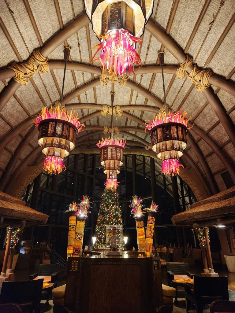 A warmly lit, tropical-themed hotel lobby at Disney’s Animal Kingdom Lodge features five chandeliers with pink and orange accents and a decorated Christmas tree in the background, creating a festive and inviting atmosphere perfect for families. - Favorite Grampy Travels