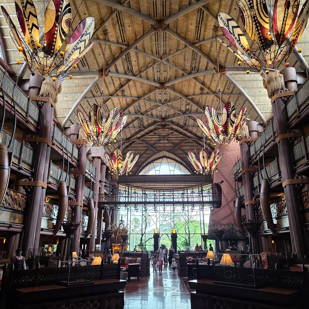 A spacious hotel lobby with tall, patterned ceilings, large decorative chandeliers, and floor-to-ceiling windows letting in natural light. Perfect for families, this inviting space resembles the grandeur of Disney’s Animal Kingdom Lodge, with people walking and sitting throughout the area—making every moment feel worth it. - Favorite Grampy Travels