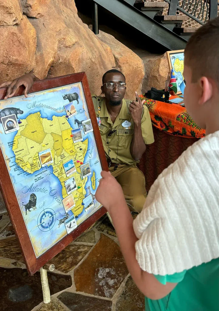 A man in uniform discusses an illustrated map of Africa with a child at Disney’s Animal Kingdom Lodge. The map features animals and landmarks, creating an engaging experience for families indoors, with rocks and a table in the background. - Favorite Grampy Travels
