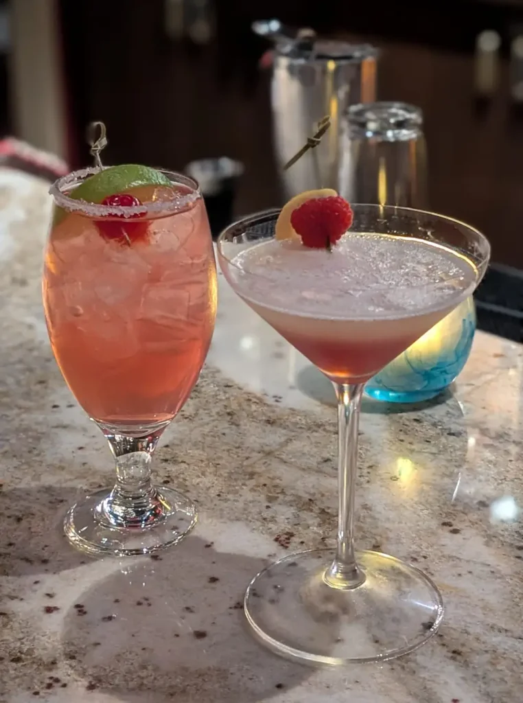 Two cocktails on a marble bar counter: one in a tall glass with ice, lime, and cherry garnish, and another in a martini glass with a frothy top, garnished with a lemon slice and raspberry—perfect for families looking to unwind after an adventurous day at Disney’s Animal Kingdom Lodge. - Favorite Grampy Travels