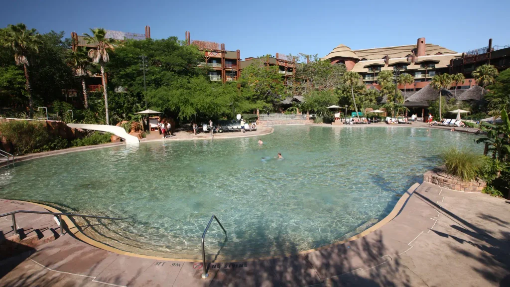 Large outdoor swimming pool with clear water surrounded by palm trees, lounge chairs, and a resort building in the background. Perfect for families, this Disney retreat offers a relaxing oasis where a few people are swimming and lounging poolside under a clear blue sky—capturing the unique atmosphere of Animal Kingdom Lodge. - Favorite Grampy Travels