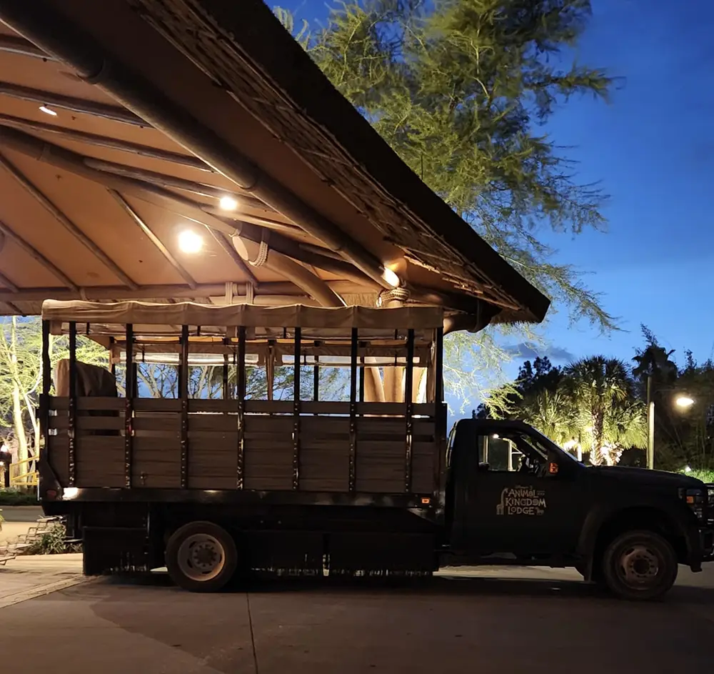 A safari truck is parked under a wooden roof at dusk at Disney’s Animal Kingdom Lodge, with warm lights illuminating the area. Families can enjoy the tranquil setting as trees and a deep blue sky form the perfect backdrop. - Favorite Grampy Travels