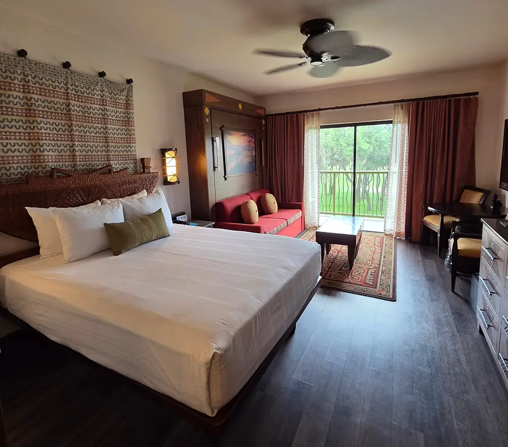 A hotel room perfect for families, featuring a large bed, red sofa, coffee table, desk, dresser, and a balcony with lush greenery outside. The room at Disney’s Animal Kingdom Lodge boasts wood floors, a ceiling fan, wall art, and abundant natural light streaming through sliding glass doors. - Favorite Grampy Travels