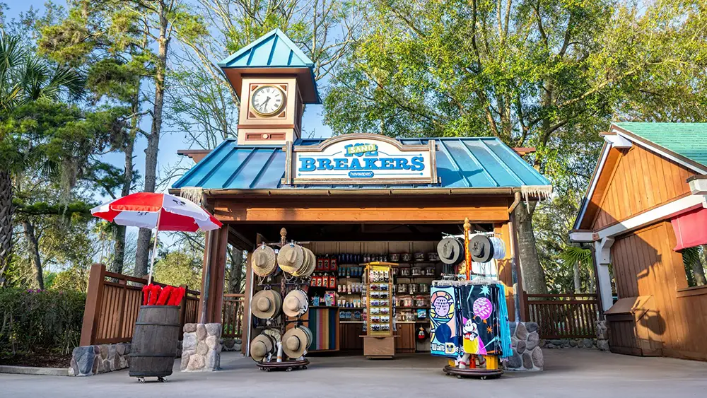 A small shop with a clock tower named Low Tide Breakers sells hats, towels, and souvenirs outdoors under a blue metal roof, surrounded by trees and a sunny sky that reflects the warm Disney World climate. - Favorite Grampy Travels