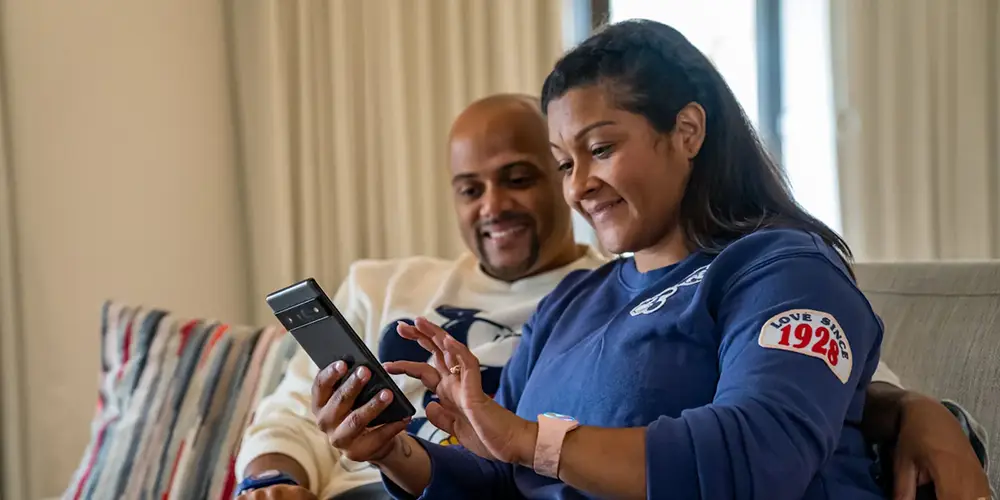 A smiling couple sits on a couch, looking at a smartphone together. The woman holds the phone, while the man sits beside her—both appearing happy and relaxed in a cozy indoor setting—as they explore Disney World Lightning Lane options for their upcoming trip. - Favorite Grampy Travels
