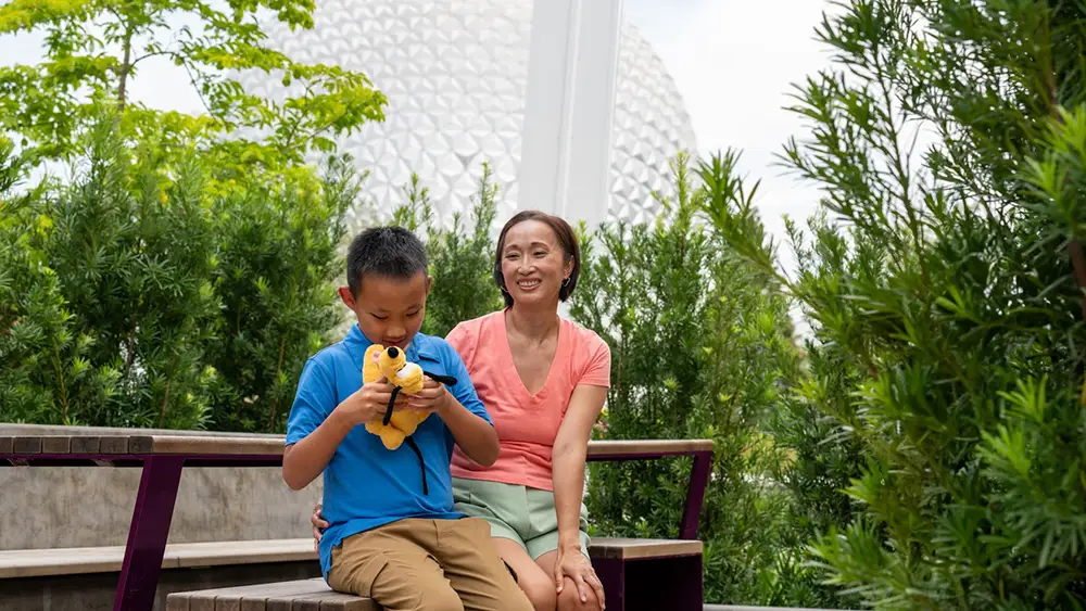 A woman and a young boy holding a Pluto plush toy sit on a wooden bench during their multi-generational Disney World trip in front of the Epcot geosphere. - Favorite Grampy Travels