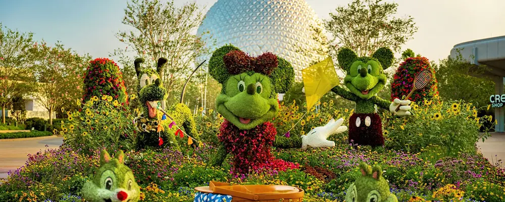 Colorful topiary sculptures of Disney characters, including Mickey, Minnie, and Goofy, are surrounded by vibrant flowers at Epcot, with the park’s iconic geodesic sphere visible in the background. Visitors can enjoy this stunning scene in any season thanks to the pleasant Disney World climate. - Favorite Grampy Travels