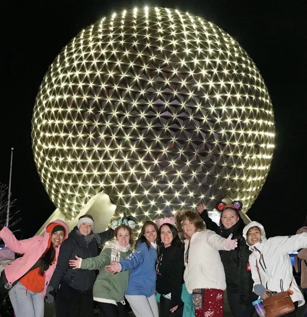 Eight people stand smiling with arms outstretched in front of EPCOT’s illuminated geodesic sphere at night, wearing winter clothes and festive accessories—a perfect snapshot for those experiencing cool evenings that are typical of Disney World weather during the winter months. - Favorite Grampy Travels