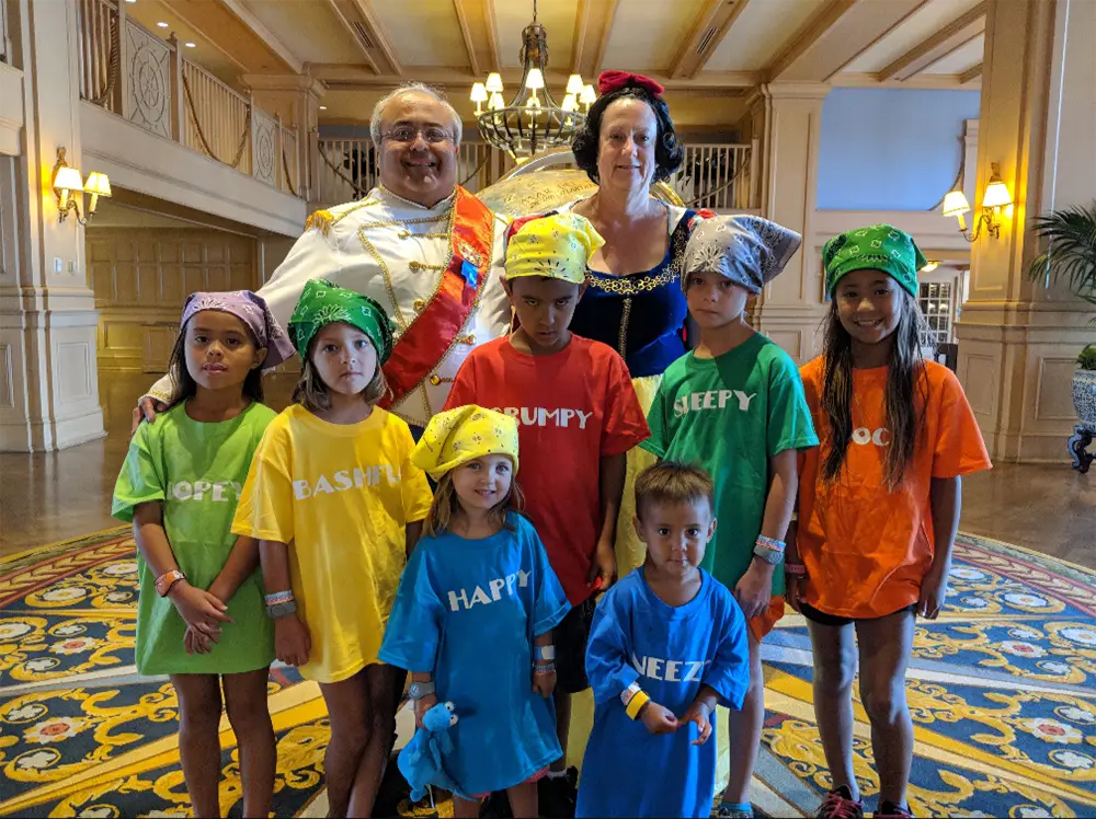A man dressed as Prince Charming and a woman dressed as Snow White stand behind seven children wearing shirts labeled with the names of the Seven Dwarfs during a multi-generational Disney trip. - Favorite Grampy Travels