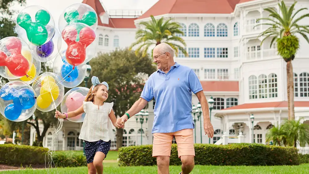 A smiling girl with Mickey Mouse balloons holds hands with an older man as they walk on grass in front of a large, white resort building with palm trees, enjoying the beautiful Disney World weather. - Favorite Grampy Travels