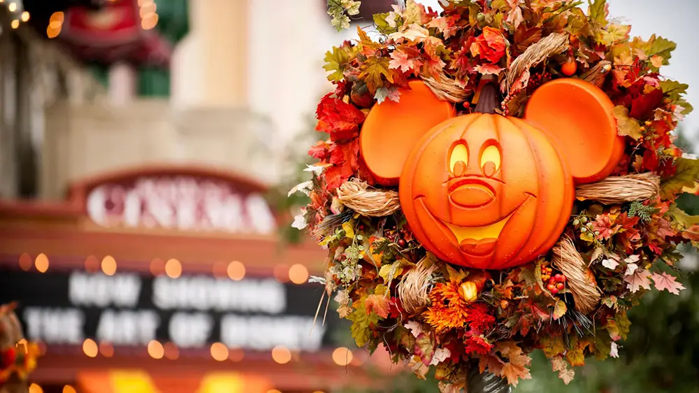 A Mickey Mouse-shaped pumpkin is surrounded by autumn leaves and flowers, set outdoors in front of a blurry theater marquee, capturing the crisp charm of Disney World’s fall weather. - Favorite Grampy Travels