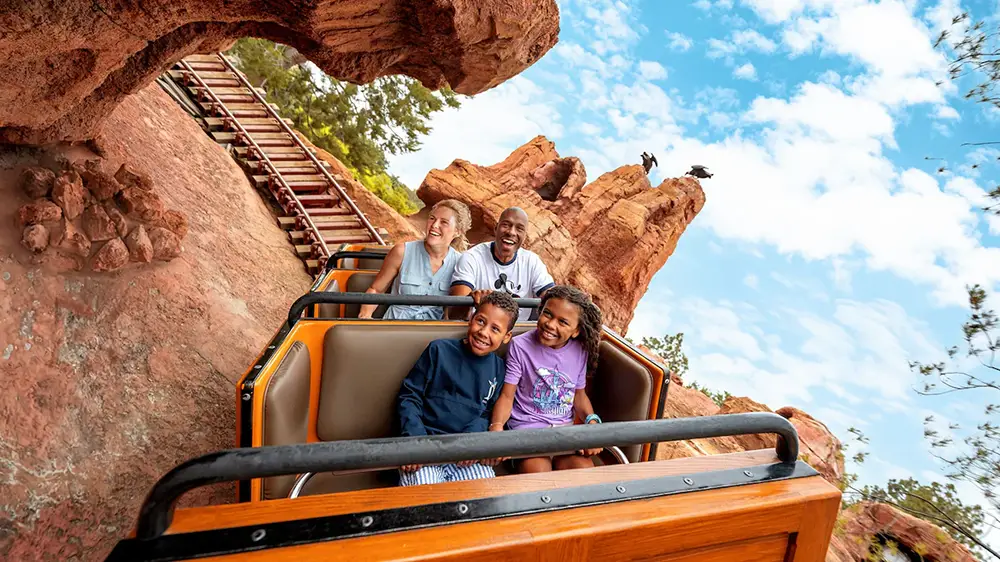 A family of four enjoys riding a roller coaster through a rocky, desert-themed landscape under a bright blue sky with scattered clouds. The children sit in front and everyone is smiling and laughing, making the most of their Disney World adventure with convenient access thanks to Lightning Lane. - Favorite Grampy Travels