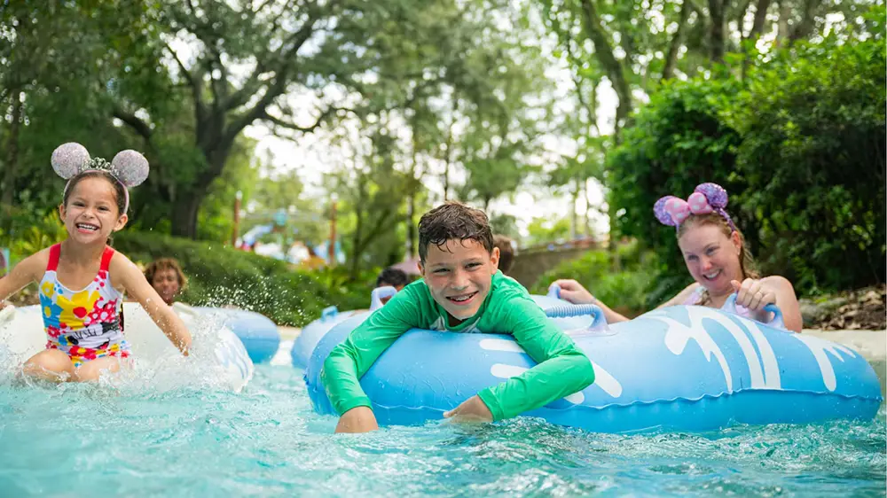 Three smiling kids wearing mouse-ear headbands enjoy a lazy river, floating on blue inner tubes and splashing water, surrounded by green trees and bright daylight—the perfect scene to experience in any season, thanks to the delightful weather at Disney World. - Favorite Grampy Travels