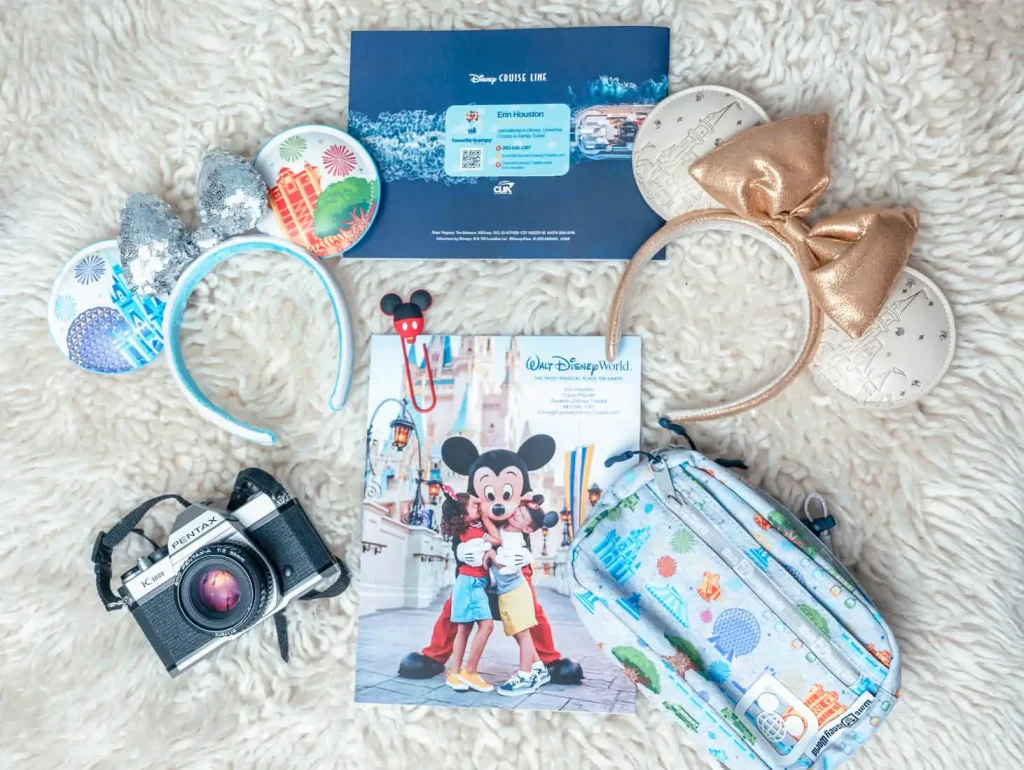 Flat lay of Disney-themed items: two Mickey Mouse ear headbands, a vintage camera, a Disney World guide with Mickey Mouse on the cover, a crossbody bag with park icons, and a blue invitation envelope. - Favorite Grampy Travels