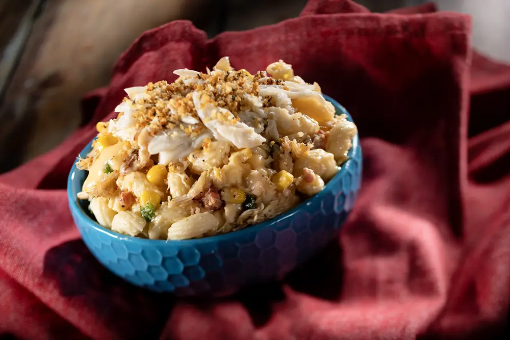 2026-wdw-food-and-wine-foodie-guide-crabmacandcheese - Favorite Grampy Travels Blue bowl of macaroni and cheese topped with crab meat and breadcrumbs on a red cloth, a popular dish during the EPCOT Food and Wine festival. - Favorite Grampy Travels