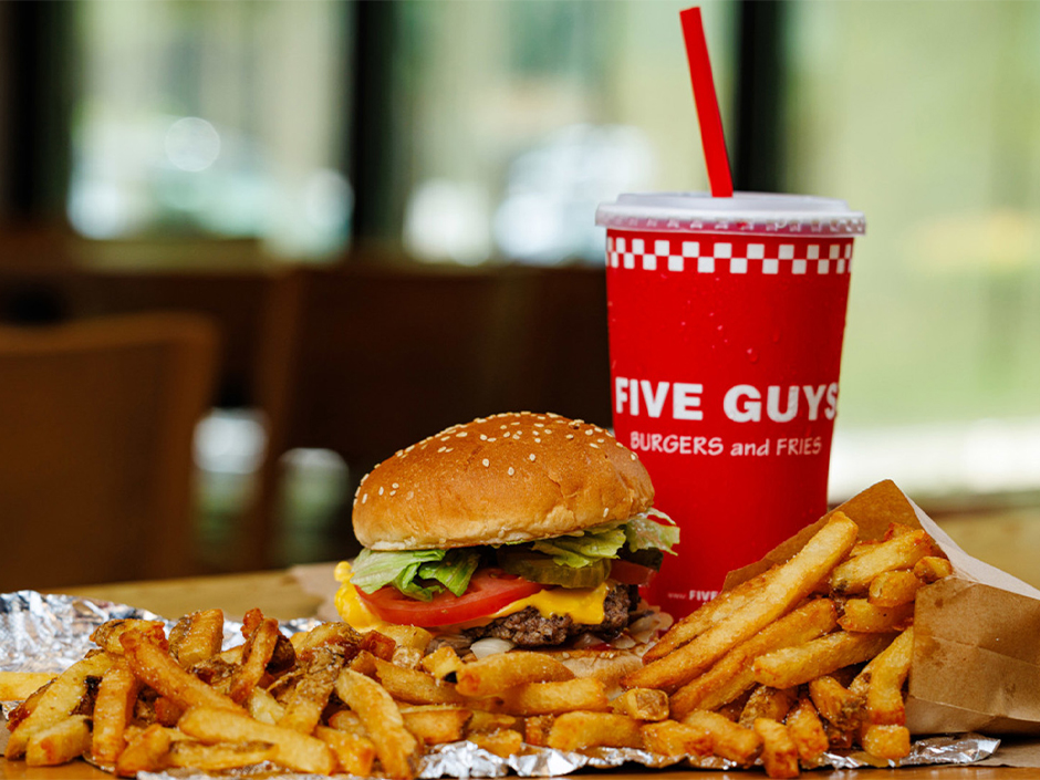 A Five Guys cheeseburger, a side of fries in a paper bag, and a red soda cup with a red straw on a wooden table, typical of a quick-service meal at Universal CityWalk. - Favorite Grampy Travels
