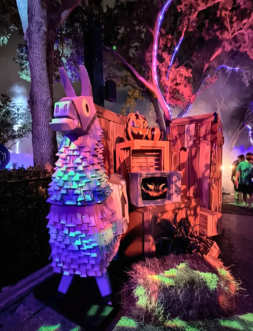 Fortnite Loot Llama Display in Fortnitemares Scare Zone at Halloween Horror Nights - Favorite Grampy Travels - Favorite Grampy Travels A purple Fortnite loot llama pinata next to a jack-o-lantern TV display in the Fortnitemares scare zone at night.