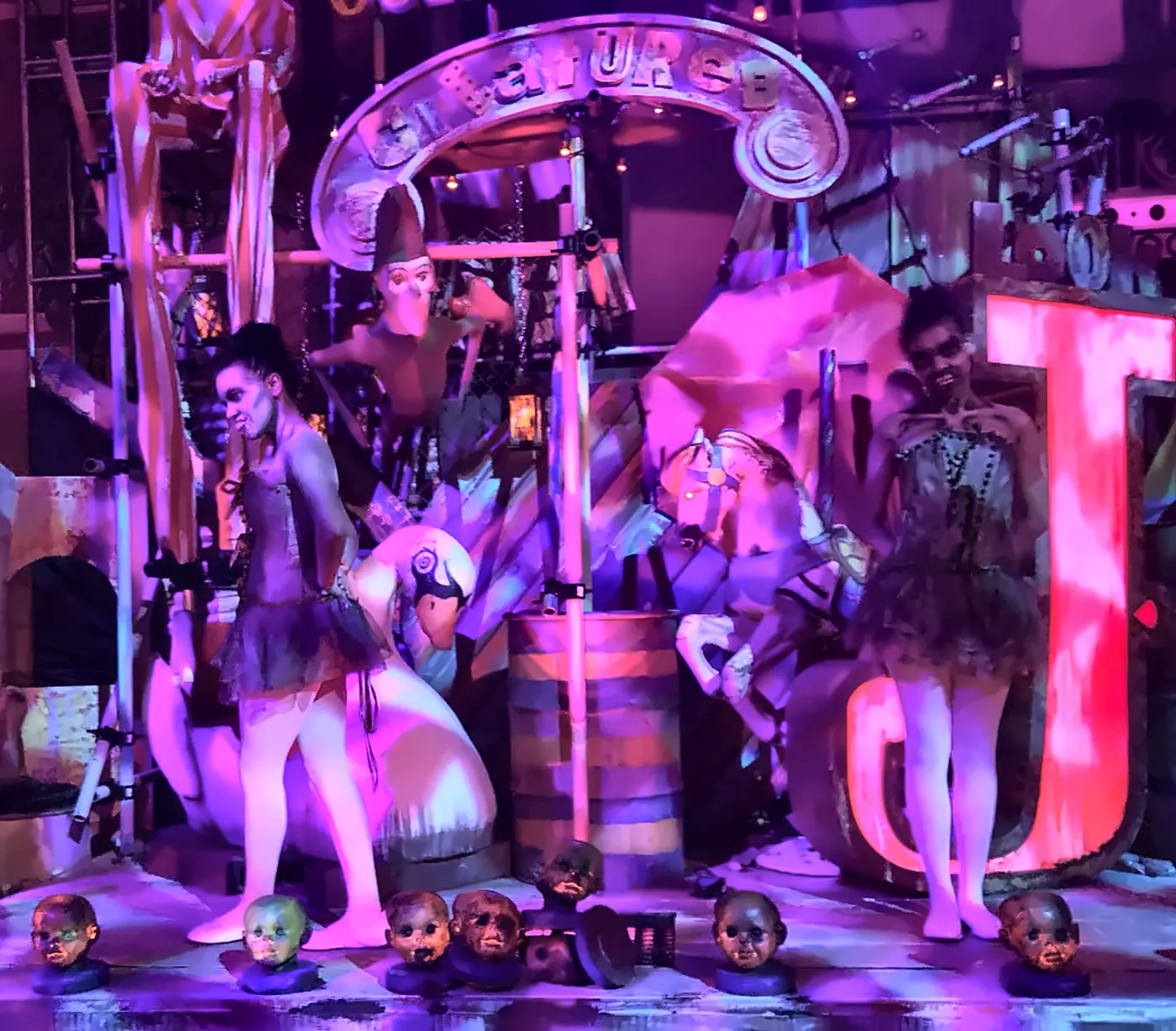 Sideshow of Decay Scare Zone Performers on Carnival Stage at Halloween Horror Nights - Favorite Grampy Travels - Favorite Grampy Travels Two performers in dark tutus posing on a carnival-themed stage with a row of weathered doll heads in the Sideshow of Decay scare zone at Halloween Horror Nights.
