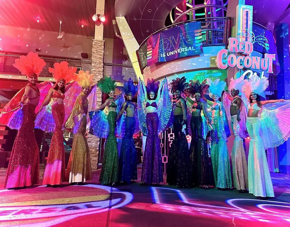 Eleven performers on stilts, wearing brightly colored sequined costumes with large wings and feathered headpieces, stand in a line at the Red Coconut Club in Universal CityWalk. - Favorite Grampy Travels