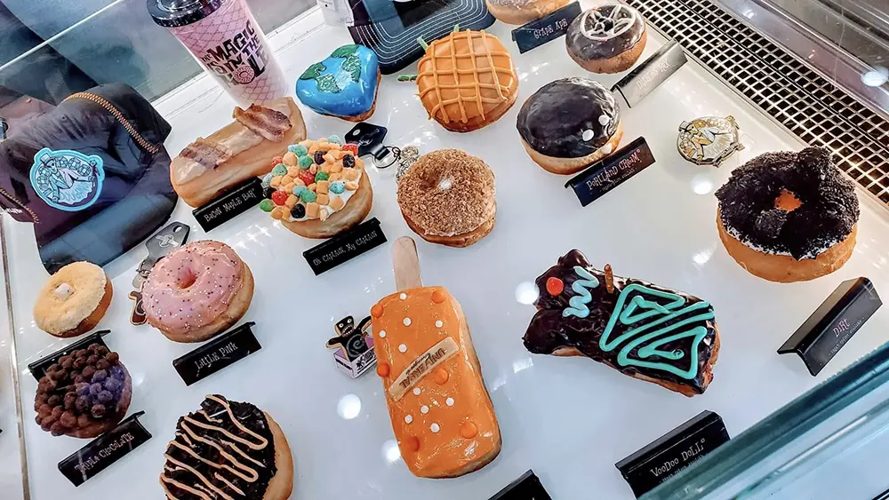 A display case of gourmet donuts at Universal CityWalk Orlando, featuring a bacon-topped donut, fruit cereal-covered treat, a rectangular donut on a stick, and a Voodoo Doll-shaped donut, each with its own descriptive label. - Favorite Grampy Travels