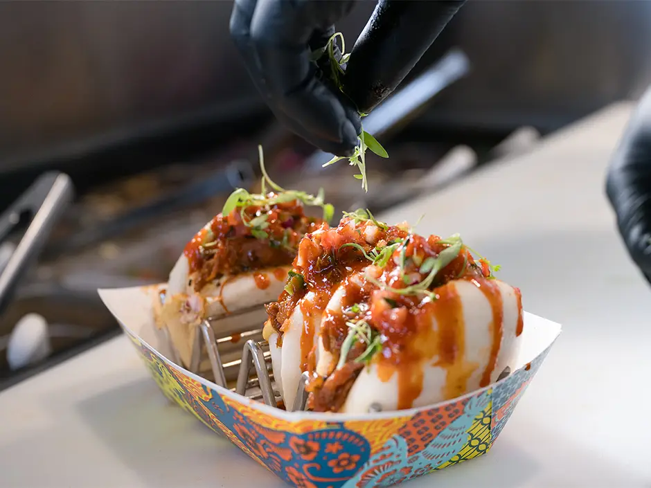 A person wearing black gloves garnishes two pulled-pork bao buns with microgreens at a Universal CityWalk quick-service meal. - Favorite Grampy Travels