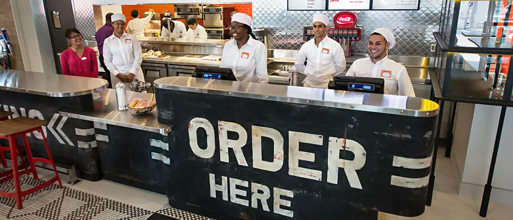 Four employees in white uniforms stand behind a restaurant counter labeled with large ORDER HERE lettering at a Universal CityWalk quick-service meal location. - Favorite Grampy Travels