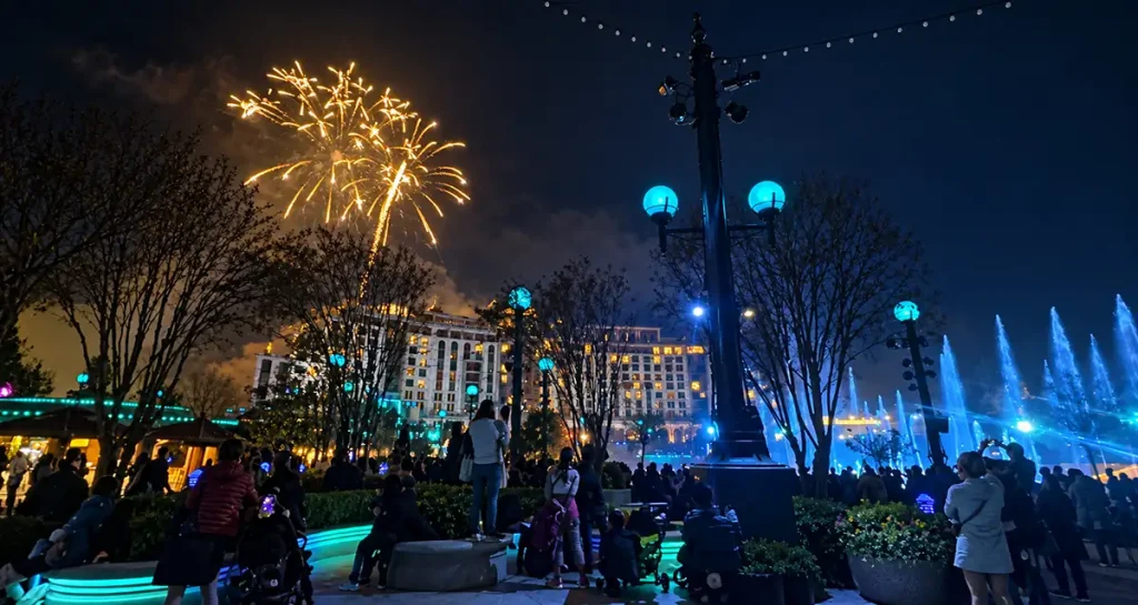 A crowd watches fireworks burst over a large hotel and illuminated fountains at night. - Favorite Grampy Travels