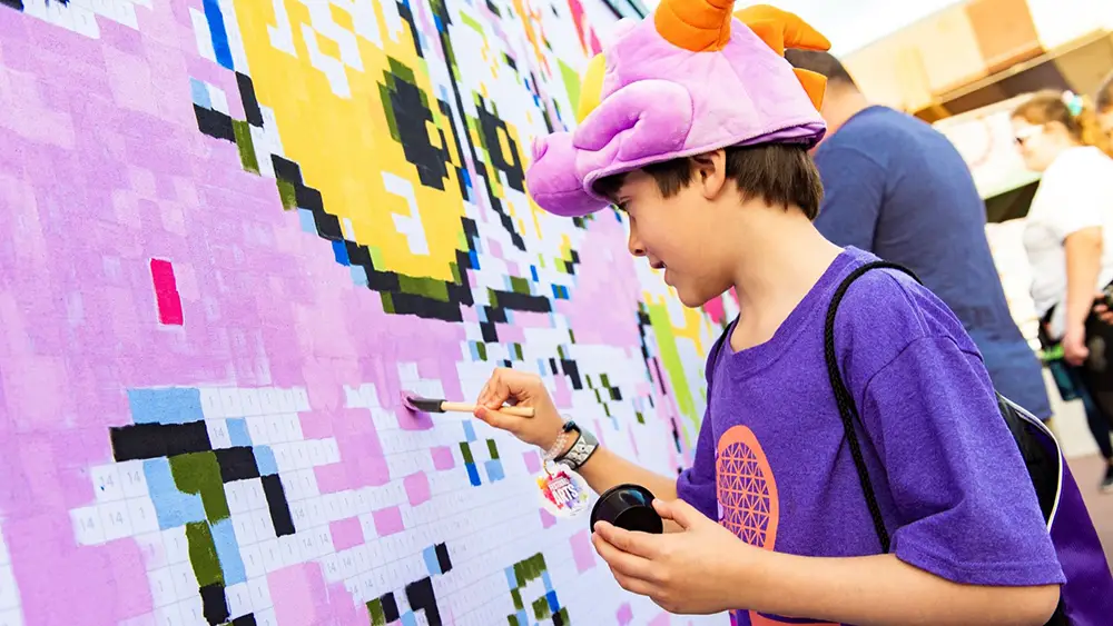 Wdw-2026-epcot-international-festival-of-the-arts-kid-painting - Favorite Grampy Travels A child wearing a purple dragon hat paints a pixel-art mural at EPCOT with kids by matching colors to numbered squares. - Favorite Grampy Travels
