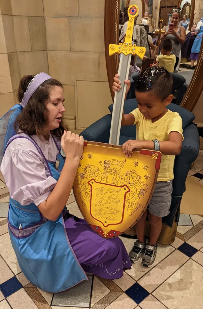 A woman in a costume at the Bibbidi Bobbidi Boutique kneeling to help a young boy hold a large toy sword and shield. - Favorite Grampy Travels