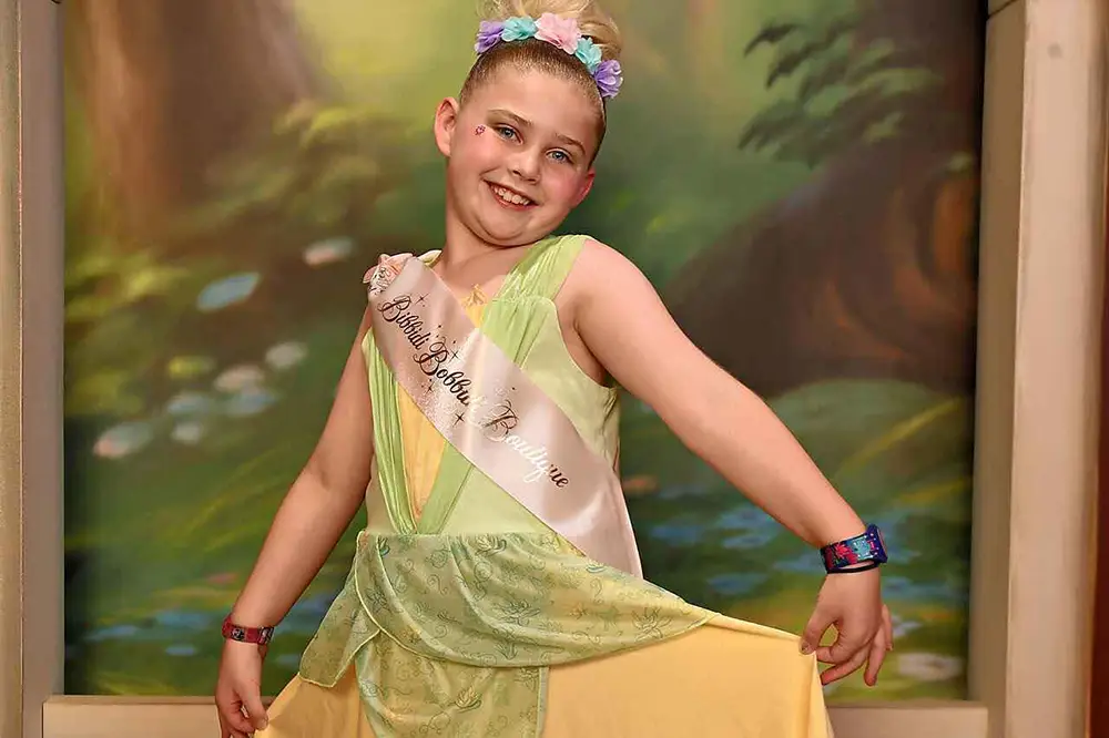 A young girl in a yellow dress and floral headband wears a Bibbidi Bobbidi Boutique sash while posing for her Disney princess makeover in front of a forest backdrop. - Favorite Grampy Travels
