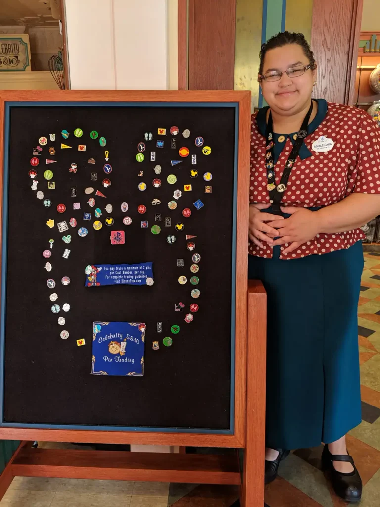 A Disney cast member stands next to a large display board filled with Mickey Mouse-shaped pins, a common sight for those interested in Disney pin trading. - Favorite Grampy Travels