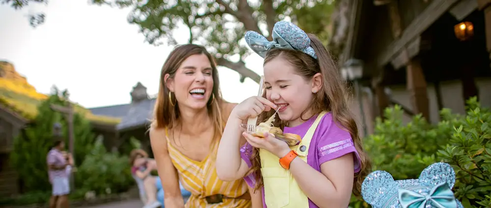 Wdw-epcot-food-and-wine-festival-emiles-fromage-montage-mom-and-daughter - Favorite Grampy Travels A young girl wearing mouse ears eats a snack at EPCOT Food and Wine while a woman behind her laughs. - Favorite Grampy Travels