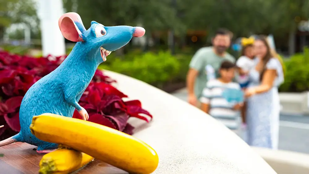 Wdw-epcot-food-and-wine-festival-remy-hide-and-squeak-with-squash - Favorite Grampy Travels A statue of Remy from Ratatouille stands next to two yellow zucchinis, with a blurred family enjoying the EPCOT Food and Wine festival in the background. - Favorite Grampy Travels