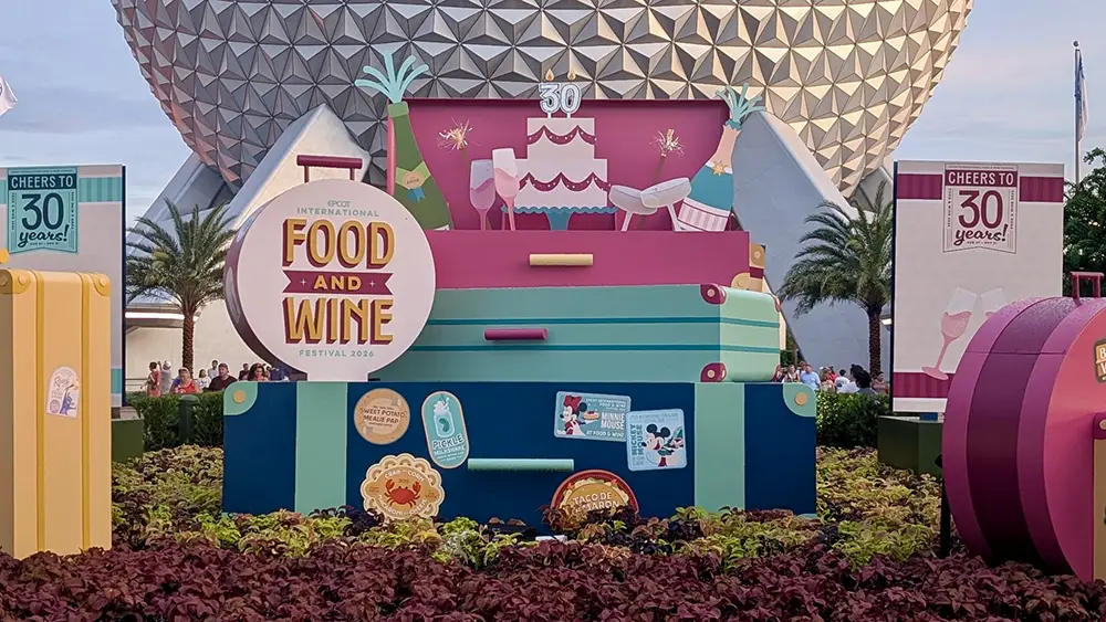 Epcot International Food and Wine Festival 2026 display with a large circular sign, a tiered cake decoration, and promotional signage for a family guide to EPCOT in front of Spaceship Earth. - Favorite Grampy Travels