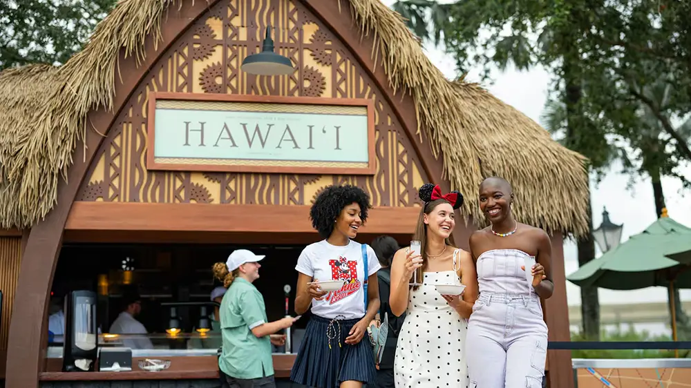 Wdw-epcot-food-and-wine-hawaii-festival-booth - Favorite Grampy Travels Three people smile while holding small bowls in front of a thatched-roof Hawaii kiosk at the EPCOT Food and Wine festival. - Favorite Grampy Travels